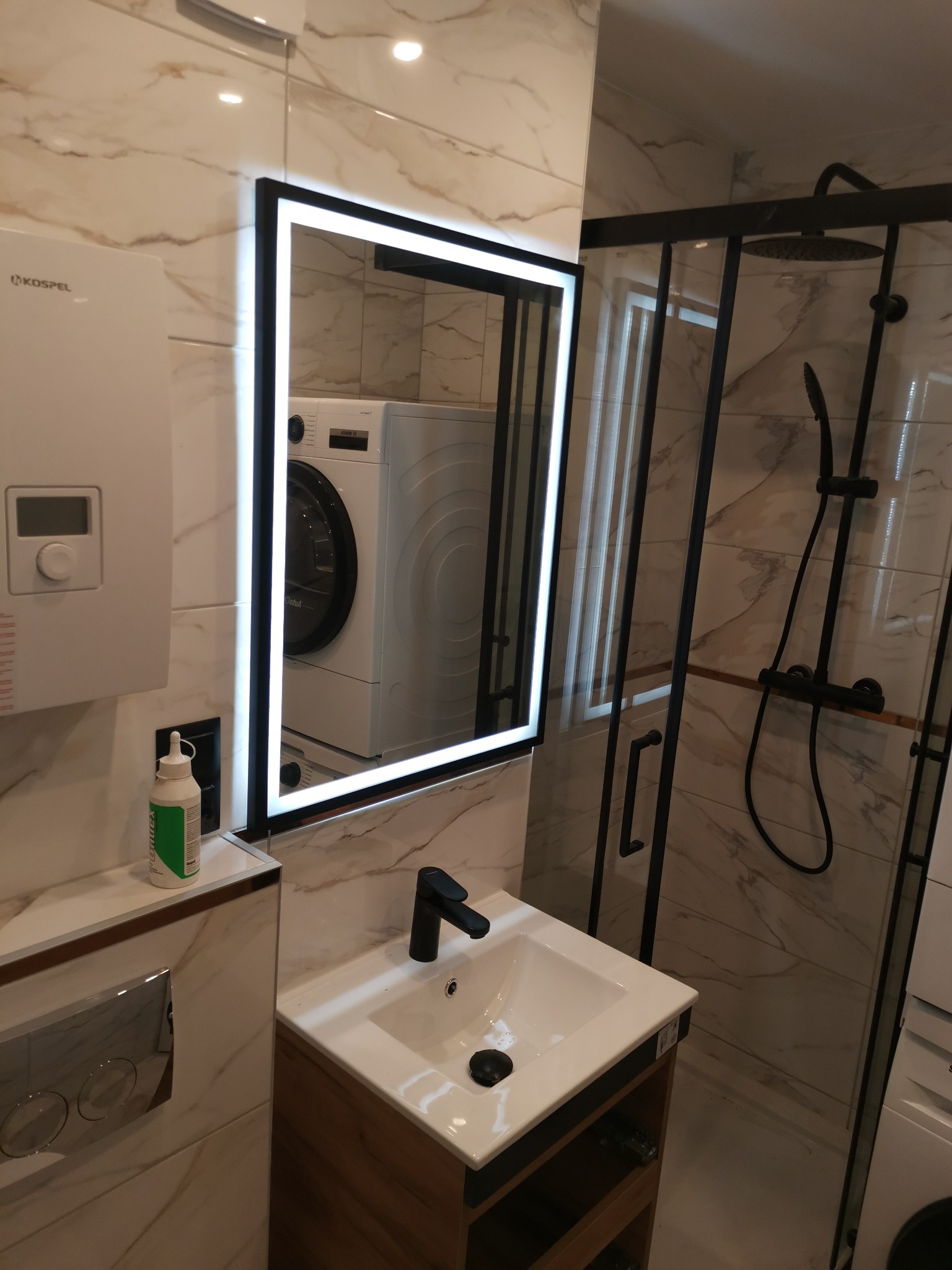 Nowoczesna łazienka z kabiną prysznicową z czarnymi profilami, umywalką z szafką z drewnopodobnym wykończeniem, lustrem z podświetleniem LED i pralką widoczną w odbiciu.