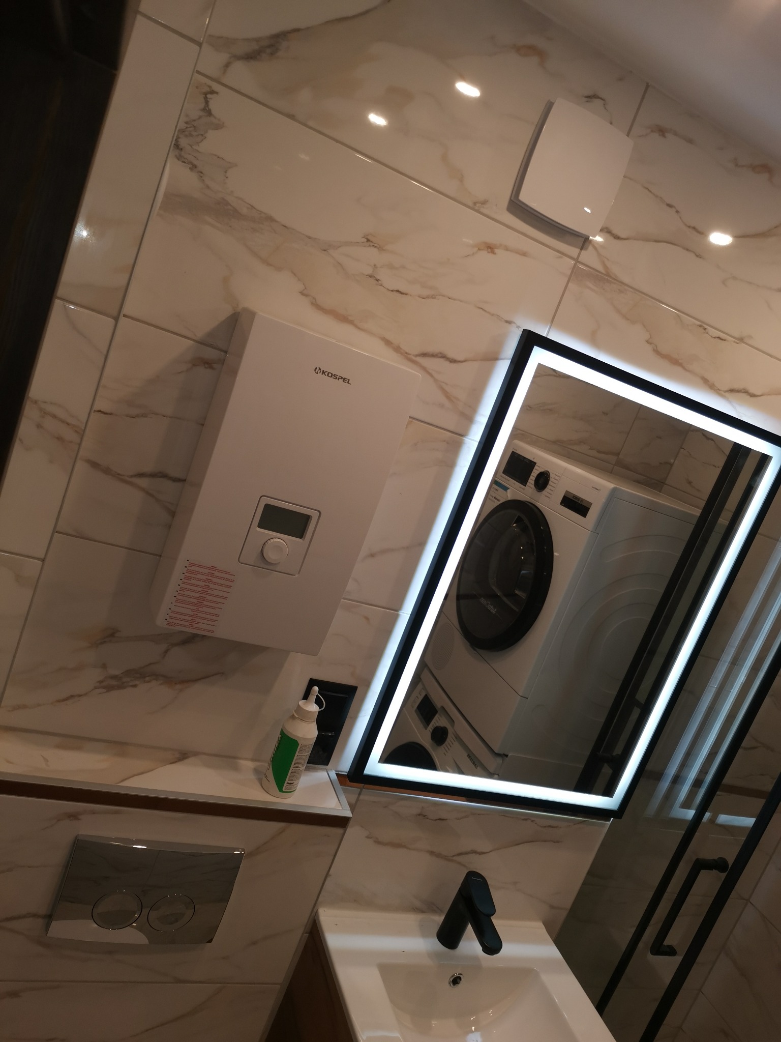 Nowoczesna łazienka z marmurowymi płytkami, podświetlanym lustrem odbijającym pralkę, bojlerem Kospel i czarną armaturą.