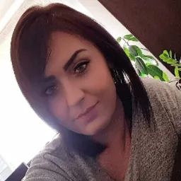 Portret kobiety z ciemnymi włosami i delikatnym makijażem, lekko pochylona głowa, w tle widoczna roślina doniczkowa.