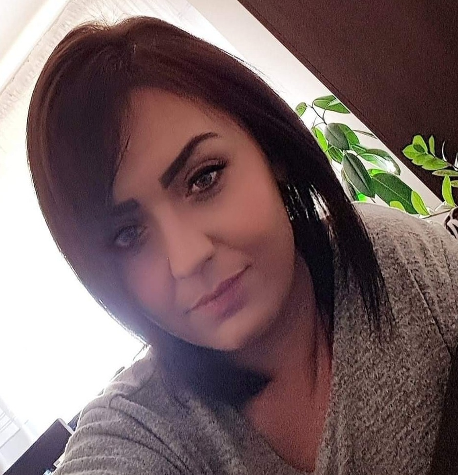 Portret kobiety z ciemnymi włosami i delikatnym makijażem, lekko pochylona głowa, w tle widoczna roślina doniczkowa.