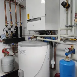 Kompaktowa kotłownia: biały wiszący piec gazowy De Dietrich, cylindryczny zasobnik ciepłej wody Remeha Aqua Plus, miedziane rury instalacji grzewczej z manometrami, białe rury PCV, zawory, filtry...