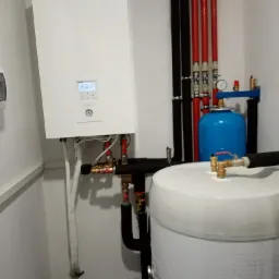 Biała pompa ciepła Panasonic Aquarea z wyświetlaczem pokazującym temperaturę 40 stopni, podłączona do instalacji z rurami w kolorach czarnym i czerwonym oraz niebieskim zbiornikiem wyrównawczym...