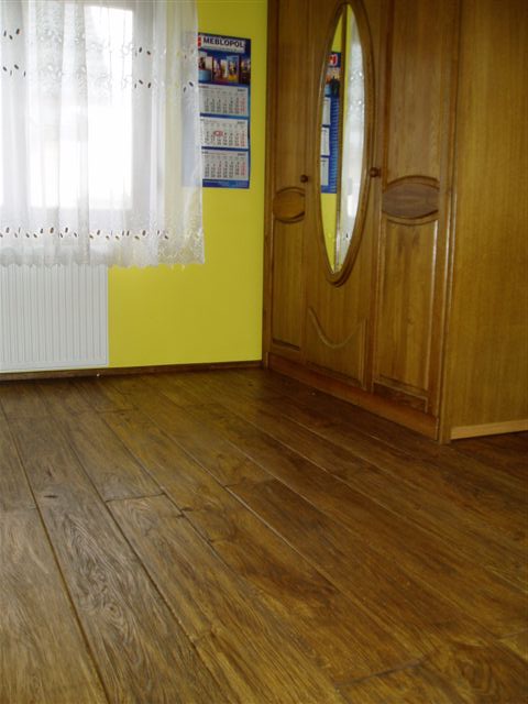 Ułożona deska podłogowa z widocznym usłojeniem, na ścianie żółta farba, obok grzejnik i szafa z owalnym lustrem.