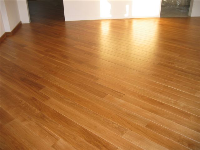 Jasna, lakierowana deska podłogowa ułożona diagonalnie, widoczne listwy przypodłogowe i fragment ściany w tle.