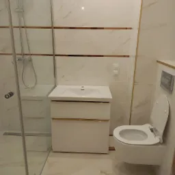 Wykończona łazienka z kabiną prysznicową ze szklanymi drzwiami, białą umywalką z szafką z złotymi akcentami, toaletą podwieszaną i płytkami imitującymi marmur z poziomymi złotymi paskami.