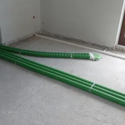 Zielone rury karbowane ułożone na surowym betonowym podłożu w trakcie instalacji w nowym budynku, widoczne także białe rurki instalacyjne przy ścianach i okno.