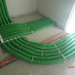 Układanie zielonych, karbowanych rur instalacyjnych na podłodze w surowym stanie, zabezpieczonych metalowymi uchwytami, obok czerwonych rur.