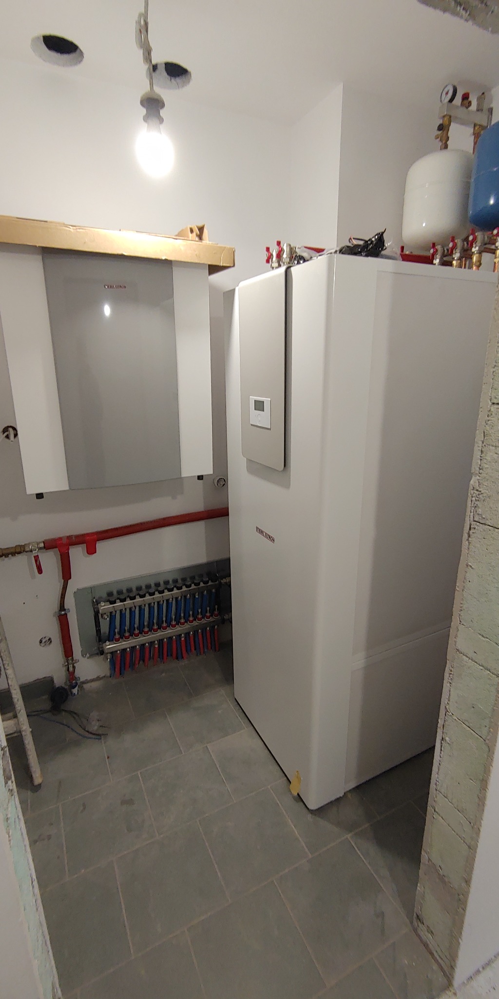 Dwa białe urządzenia grzewcze marki Stiebel Eltron zamontowane w pomieszczeniu z szarą podłogą z płytek, widoczne rury instalacji grzewczej i rozdzielacz.