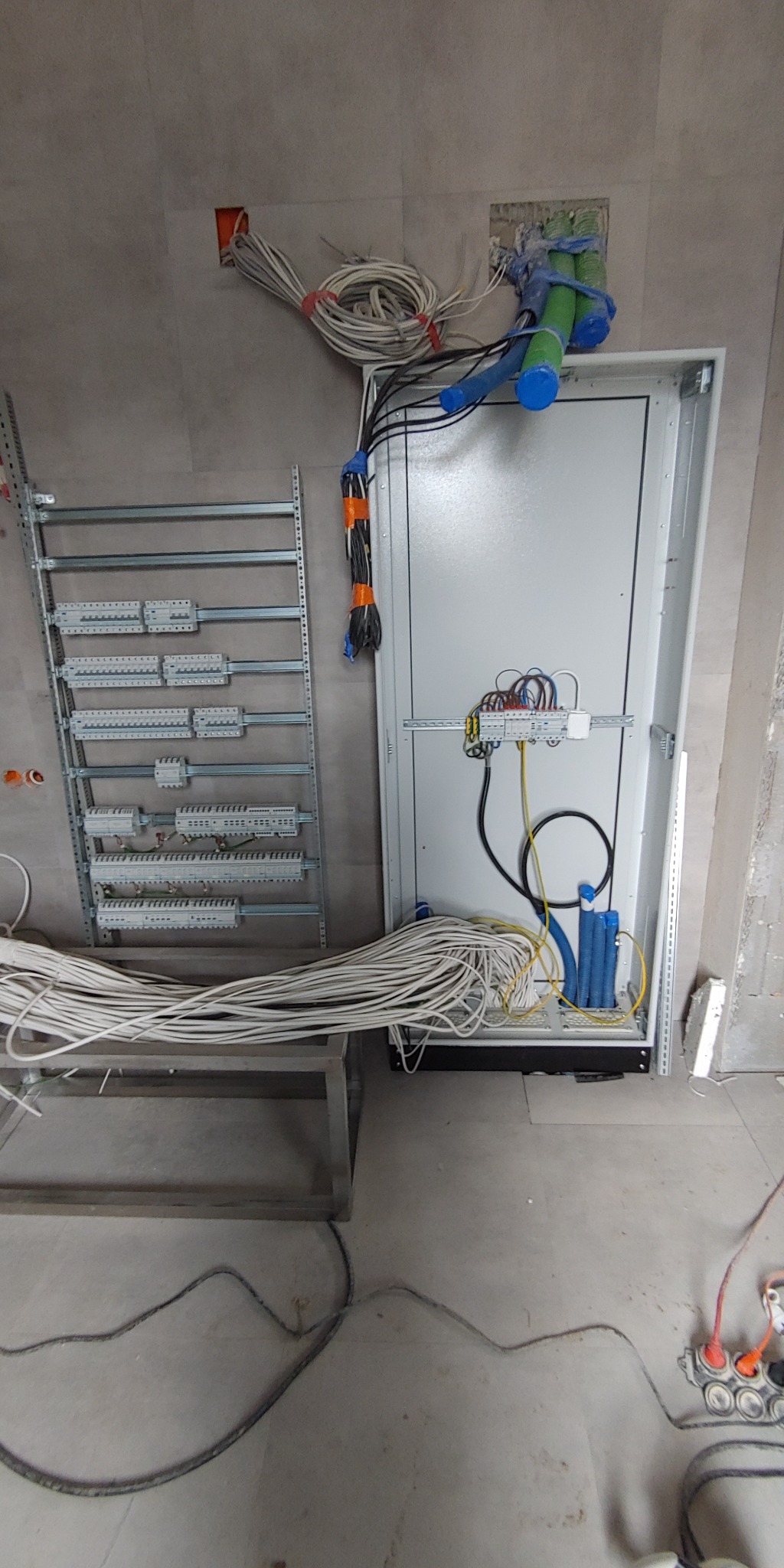 Rozdzielnica elektryczna w trakcie montażu, widoczne przewody, kable i rury instalacyjne. Ściany w szarym kolorze, surowy stan wykończenia wnętrza.