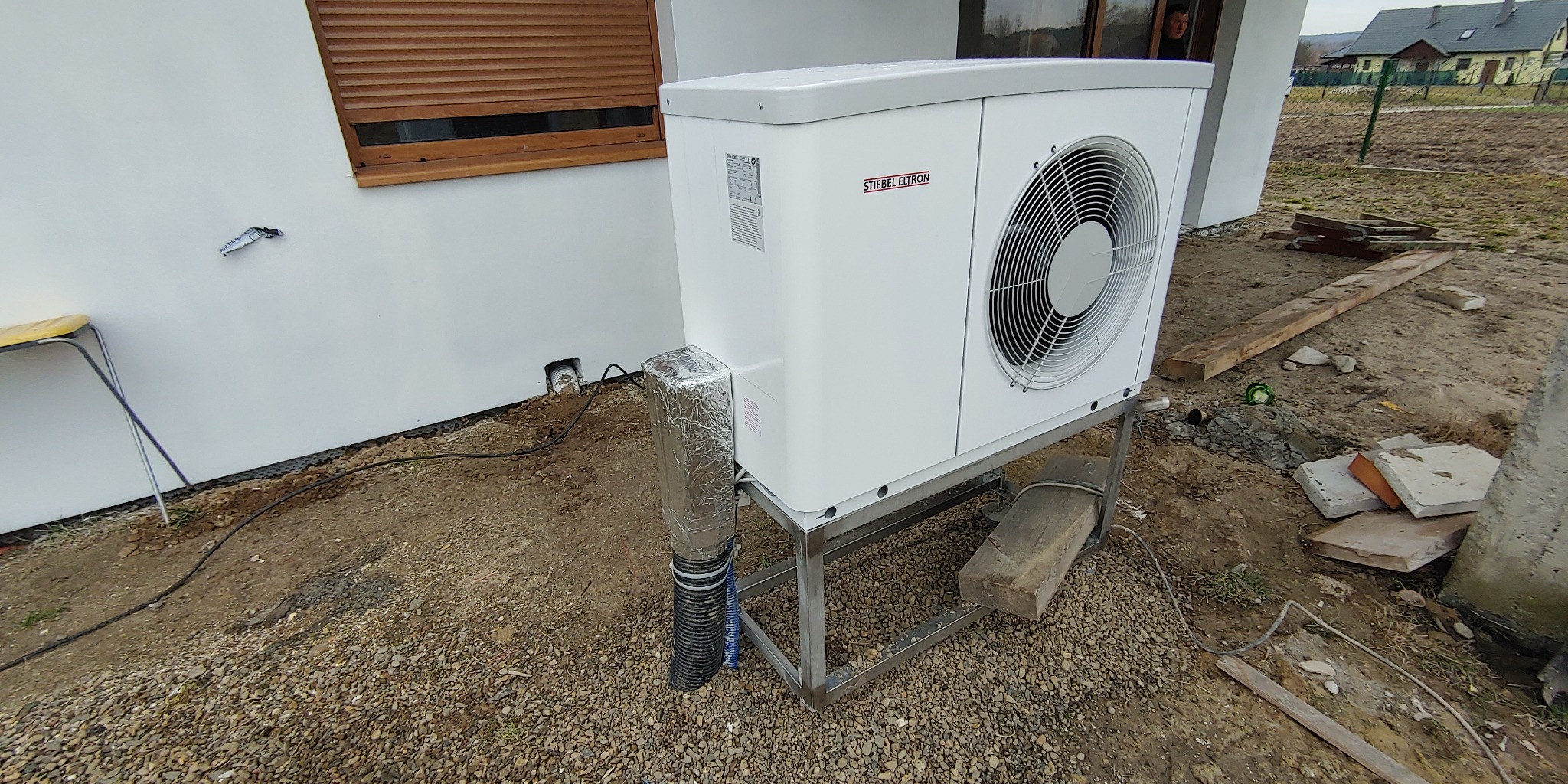 Biała pompa ciepła marki Stiebel Eltron zamontowana na metalowym stelażu przy białej ścianie budynku z brązowymi roletami, widoczne elementy izolacji rur i niezagospodarowany teren wokół.
