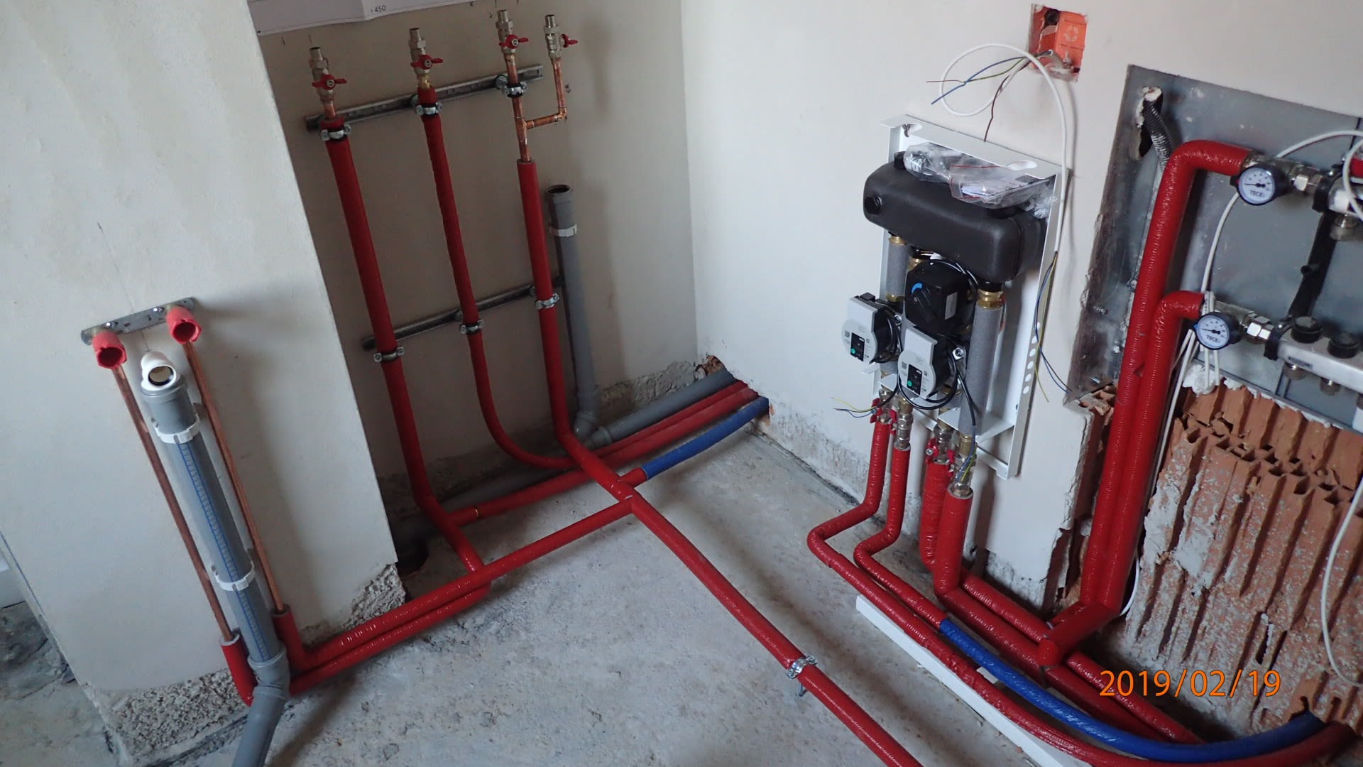 Rozbudowana instalacja centralnego ogrzewania w trakcie montażu, widoczne czerwone i niebieskie rury, rozdzielacze oraz elementy kotłowni w surowym pomieszczeniu.
