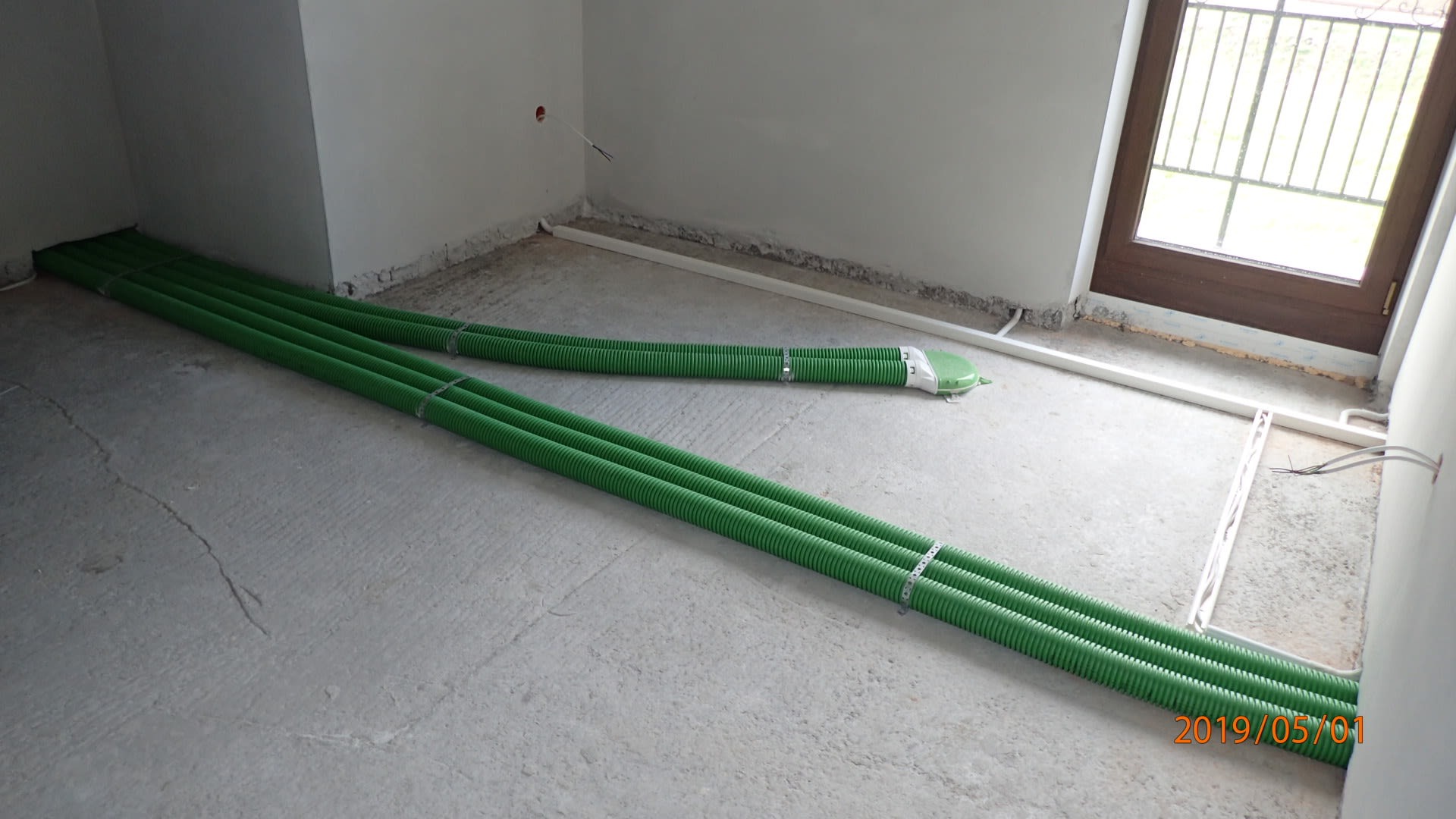 Zielone rury karbowane ułożone na surowym betonowym podłożu w trakcie instalacji w nowym budynku, widoczne także białe rurki instalacyjne przy ścianach i okno.