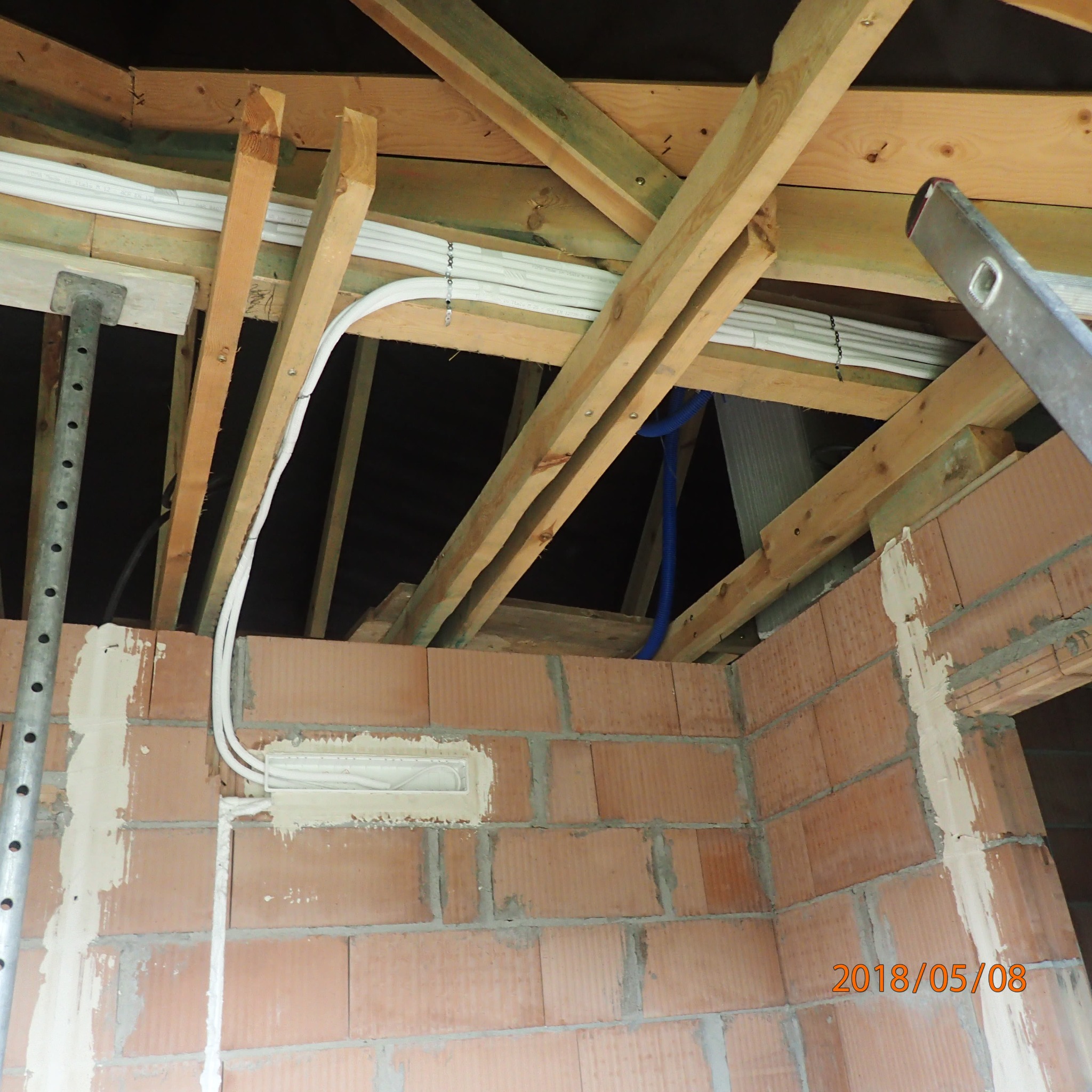 Wnętrze w trakcie budowy: widoczne drewniane elementy konstrukcyjne dachu, instalacja elektryczna w peszlach i skrzynka elektryczna wmurowana w ścianę z czerwonej cegły, fragment drabiny oparty...