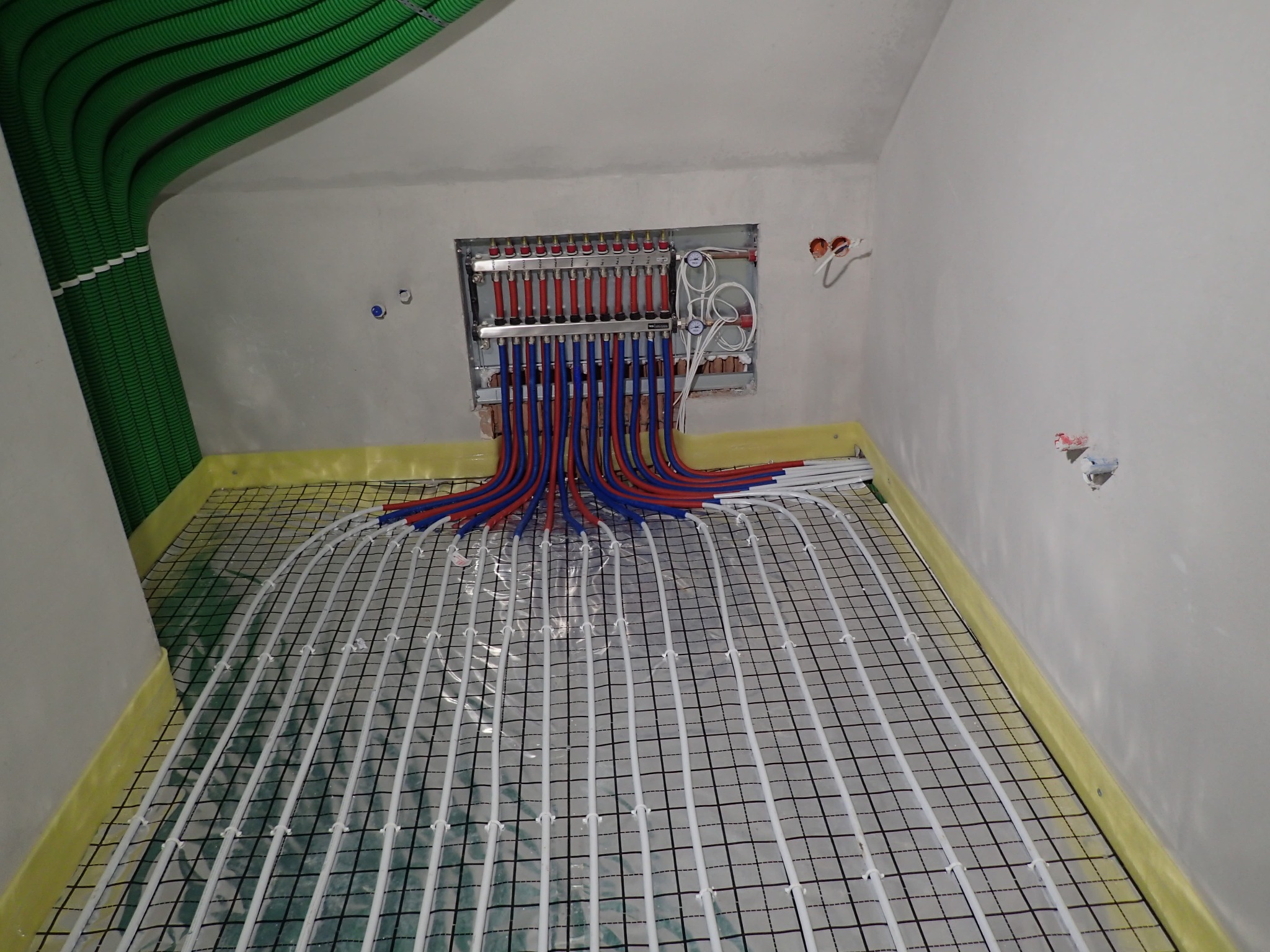 Rozłożona instalacja ogrzewania podłogowego z rurkami na siatce, widoczny rozdzielacz z rurami doprowadzającymi w kolorze czerwonym i niebieskim oraz zielone rury wentylacyjne pod sufitem w surowym...