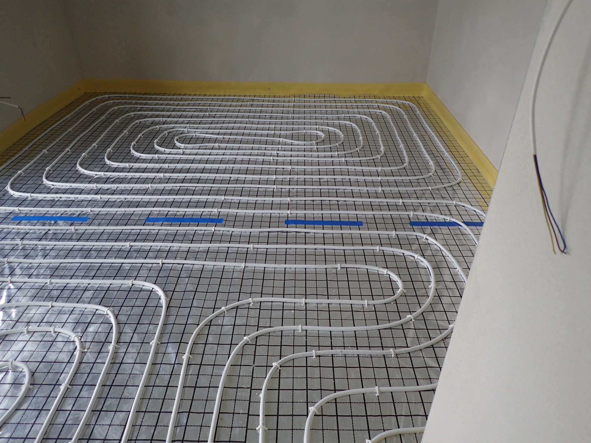 Białe rury ogrzewania podłogowego ułożone spiralnie na czarnej macie z siatką, przymocowane niebieską taśmą do żółtej izolacji przy ścianie.