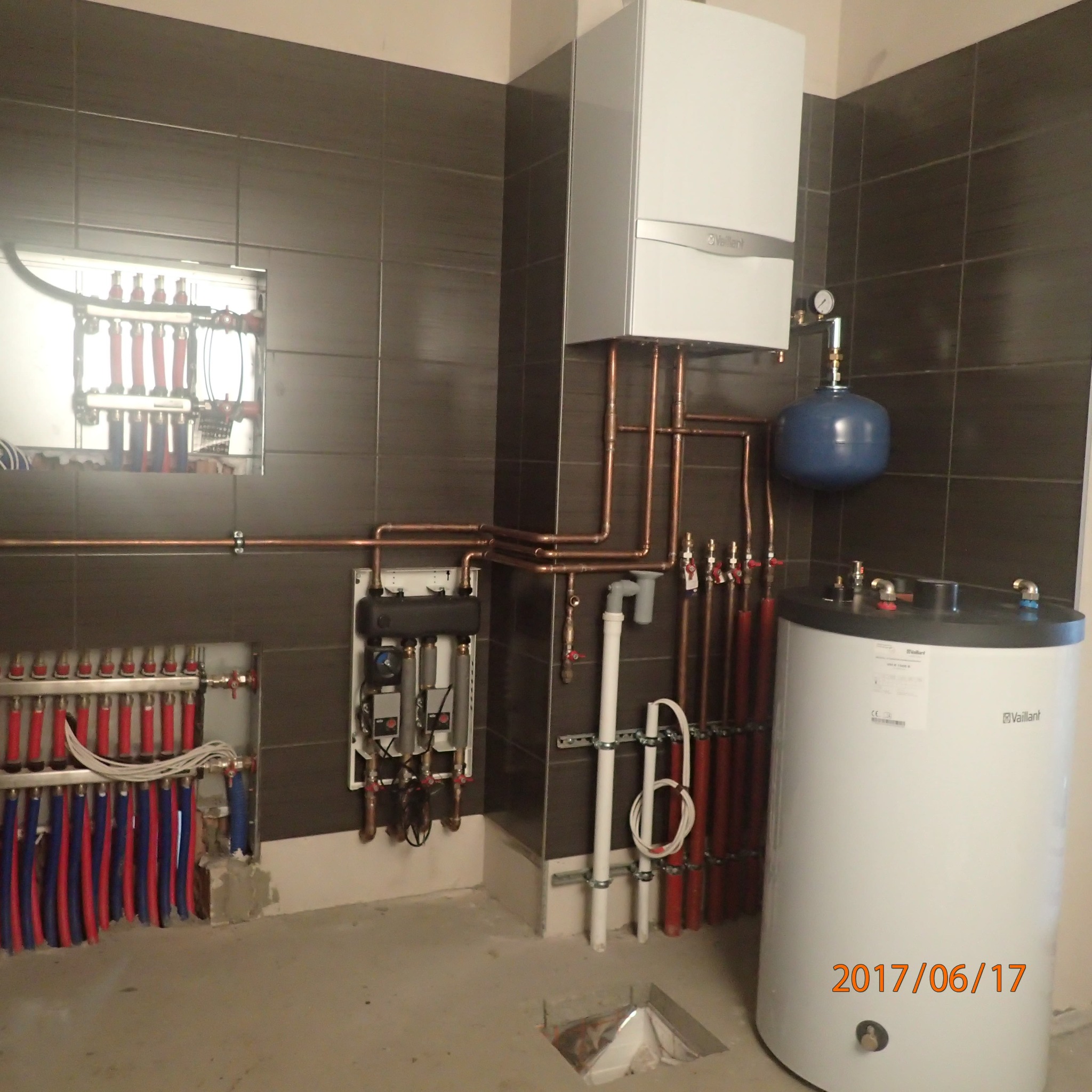 Nowoczesna kotłownia z białym zasobnikiem ciepłej wody, kotłem gazowym Vaillant, rozdzielaczami podłogowymi i miedzianą instalacją grzewczą na tle ciemnych płytek.