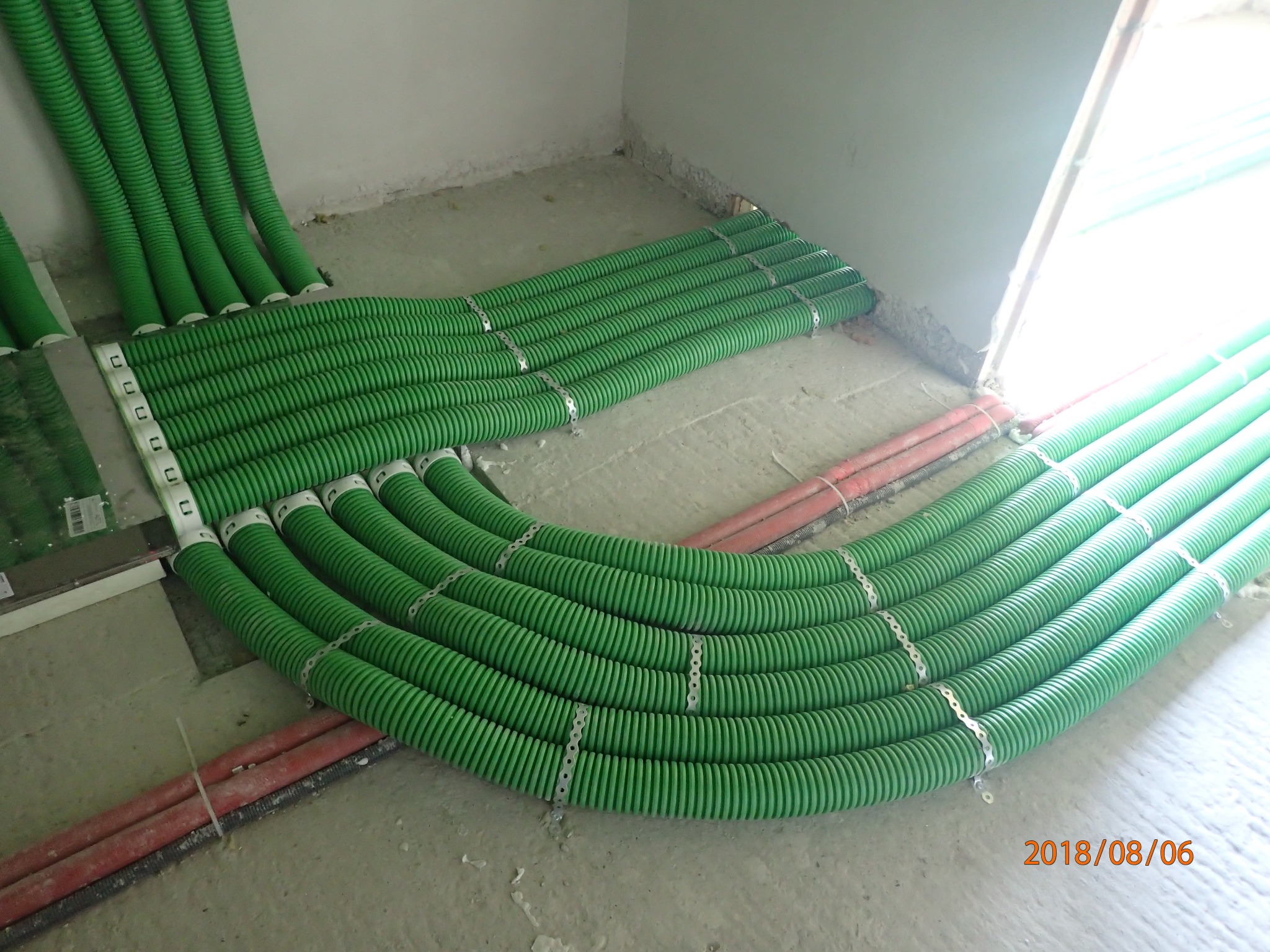 Układanie zielonych, karbowanych rur instalacyjnych na podłodze w surowym stanie, zabezpieczonych metalowymi uchwytami, obok czerwonych rur.