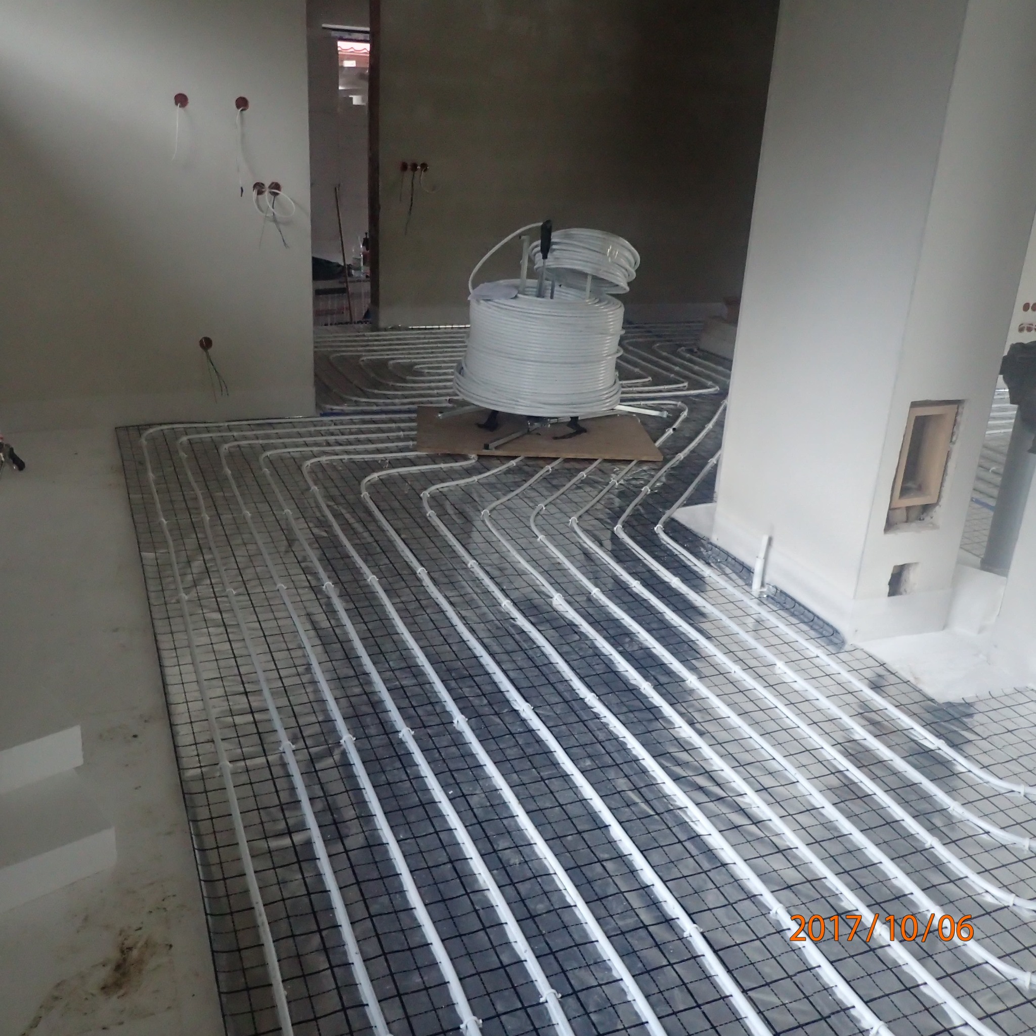 Instalacja ogrzewania podłogowego: białe rury ułożone na czarnej siatce, w tle zwój rur na tekturze, surowy stan wykończenia wnętrza.