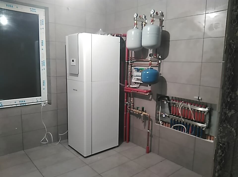 Biała pompa ciepła obok okna i rozdzielacza ogrzewania podłogowego z czerwonymi i niebieskimi rurkami w szarej, wykafelkowanej kotłowni.