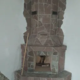 Ozdobny kominek z kamienia naturalnego w odcieniach czerwieni i szarości, z paleniskiem wypełnionym drewnem, na tle białej ściany, z widocznym kijem opartym o bok kominka.