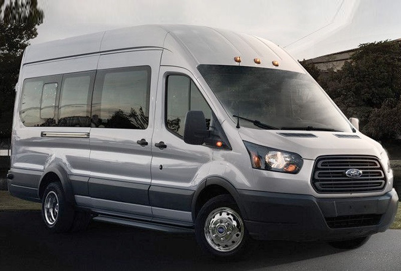 Srebrny Ford Transit z wysokim dachem i przyciemnianymi szybami, zaparkowany na asfalcie z widocznym budynkiem w tle.