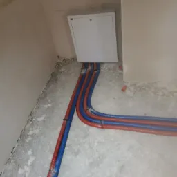 Nowe rury grzewcze (czerwone) i wodociągowe (niebieskie) prowadzone wzdłuż ściany w surowym stanie, z widoczną białą skrzynką inspekcyjną.