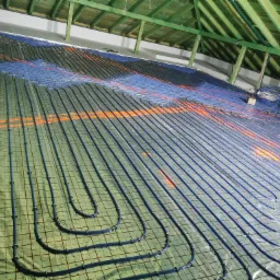 Montaż ogrzewania podłogowego: widok z góry na rozłożone rury grzewcze na folii z siatką, w tle surowa konstrukcja drewniana dachu.