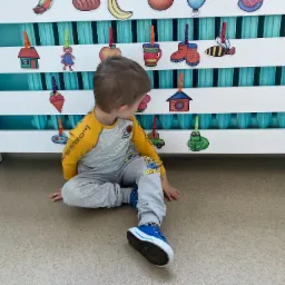 Chłopiec siedzi na podłodze, obok biały grzejnik z przyczepionymi obrazkami owoców i przedmiotów za pomocą kolorowych klamerek.