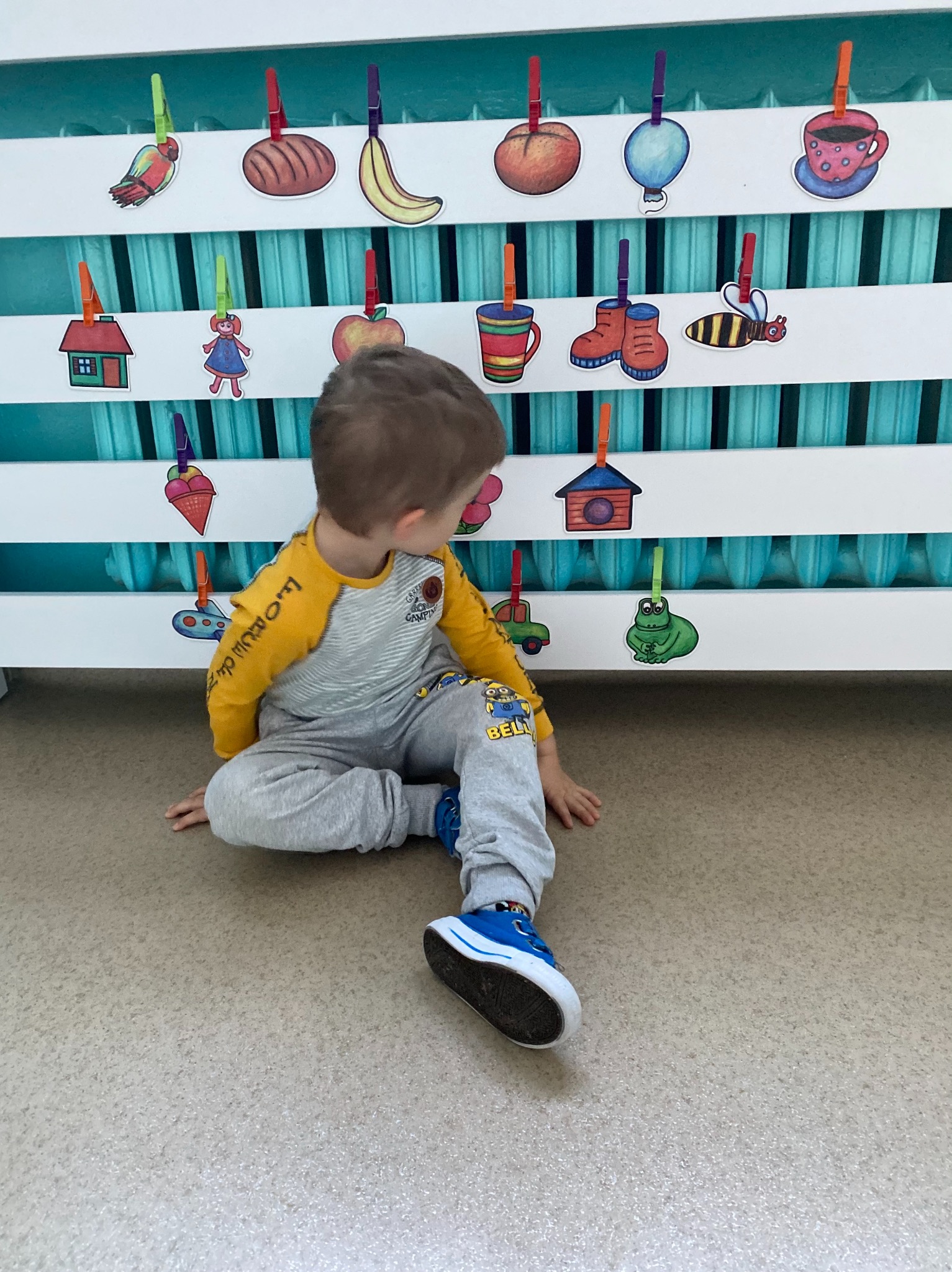 Chłopiec siedzi na podłodze, obok biały grzejnik z przyczepionymi obrazkami owoców i przedmiotów za pomocą kolorowych klamerek.