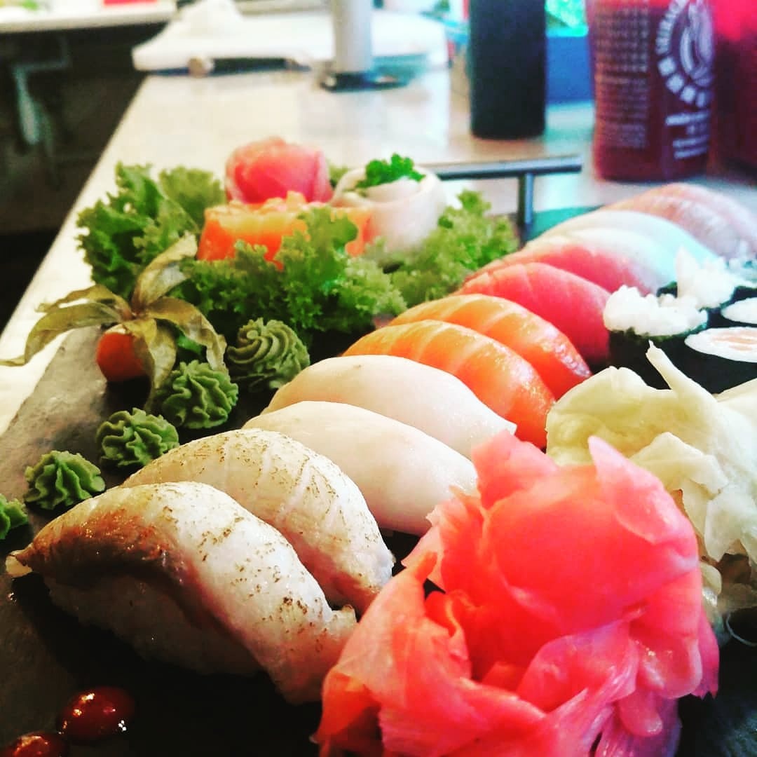 Zestaw sushi z różnymi rodzajami ryb, wasabi, imbirem i dekoracjami z sałaty na czarnym kamiennym talerzu, z butelkami sosu w tle.