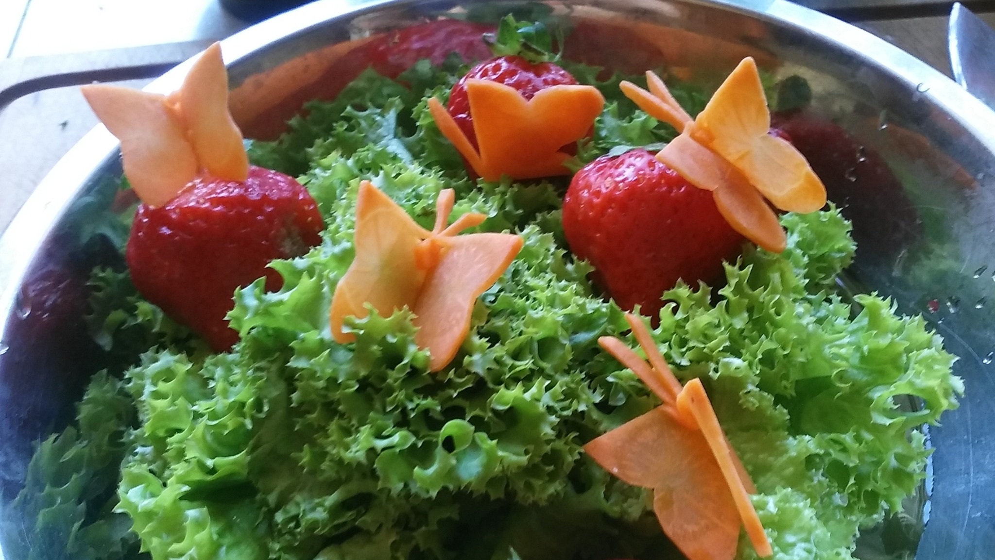 Metalowa misa z sałatą, truskawkami i dekoracjami z marchwi w kształcie motyli i kwiatów.