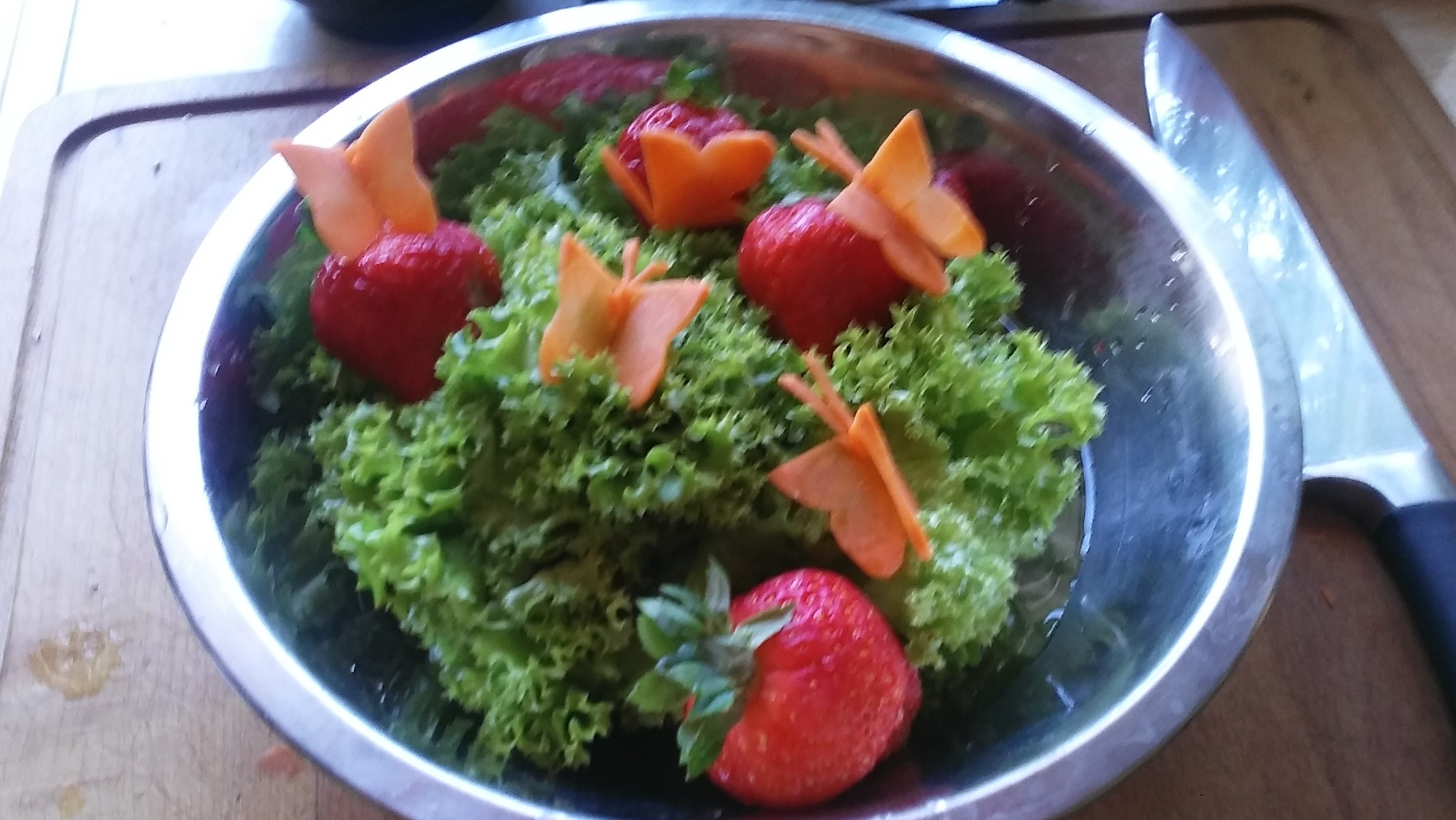 Metalowa miska z sałatą, truskawkami i dekoracjami z marchewki w kształcie motyli i kwiatów, obok nóż na drewnianej desce.