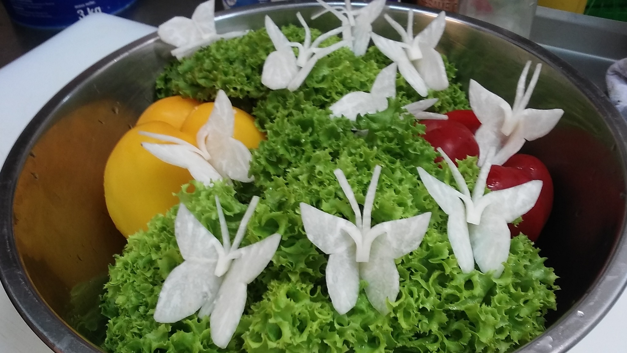 Metalowa misa z sałatą lodową, papryką żółtą i czerwoną, udekorowana rzodkiewkowymi motylami, przygotowana do cateringu.