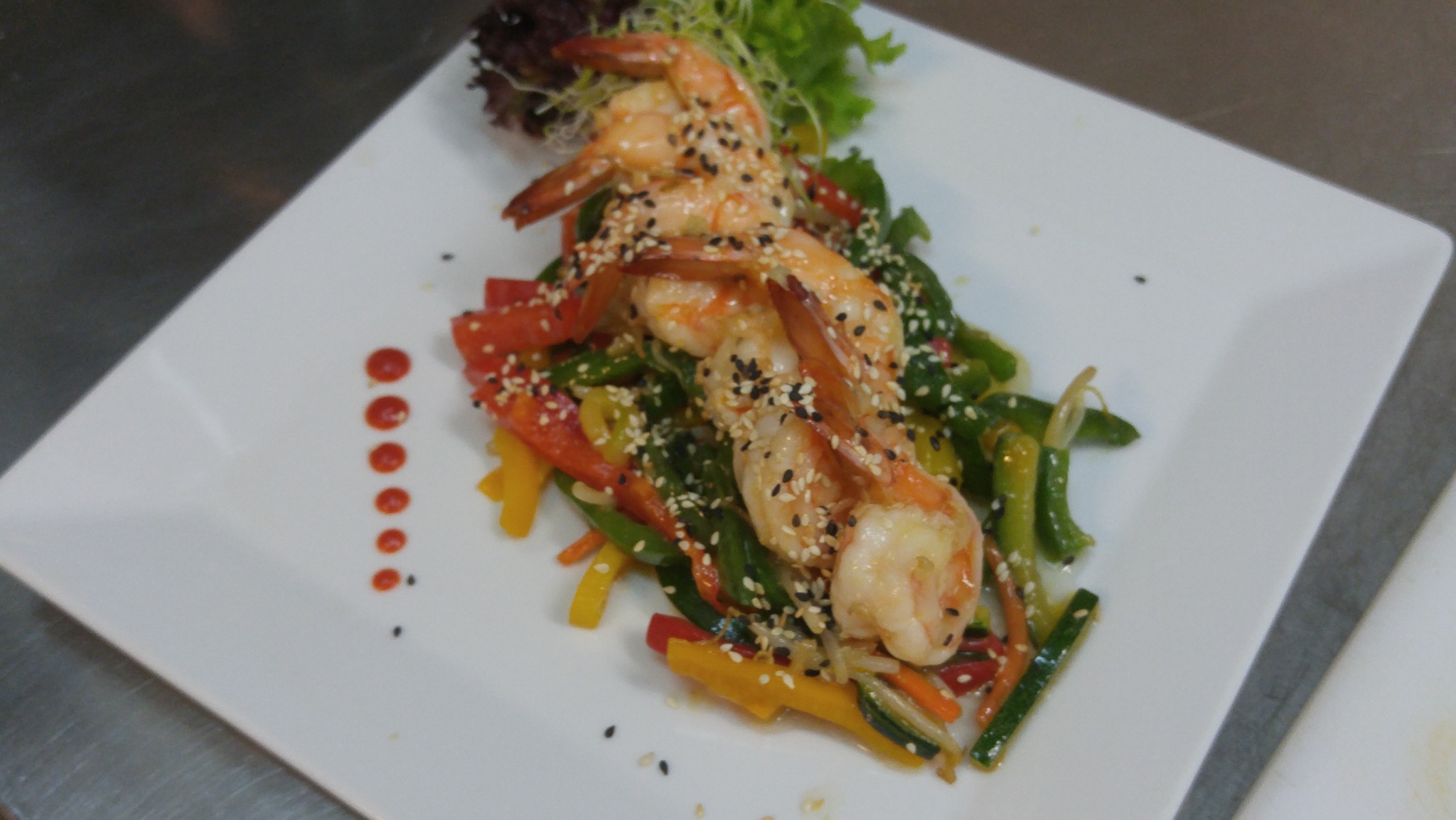 Elegancko podane krewetki z warzywami i sezamem na białym talerzu, ozdobione kroplami sosu.