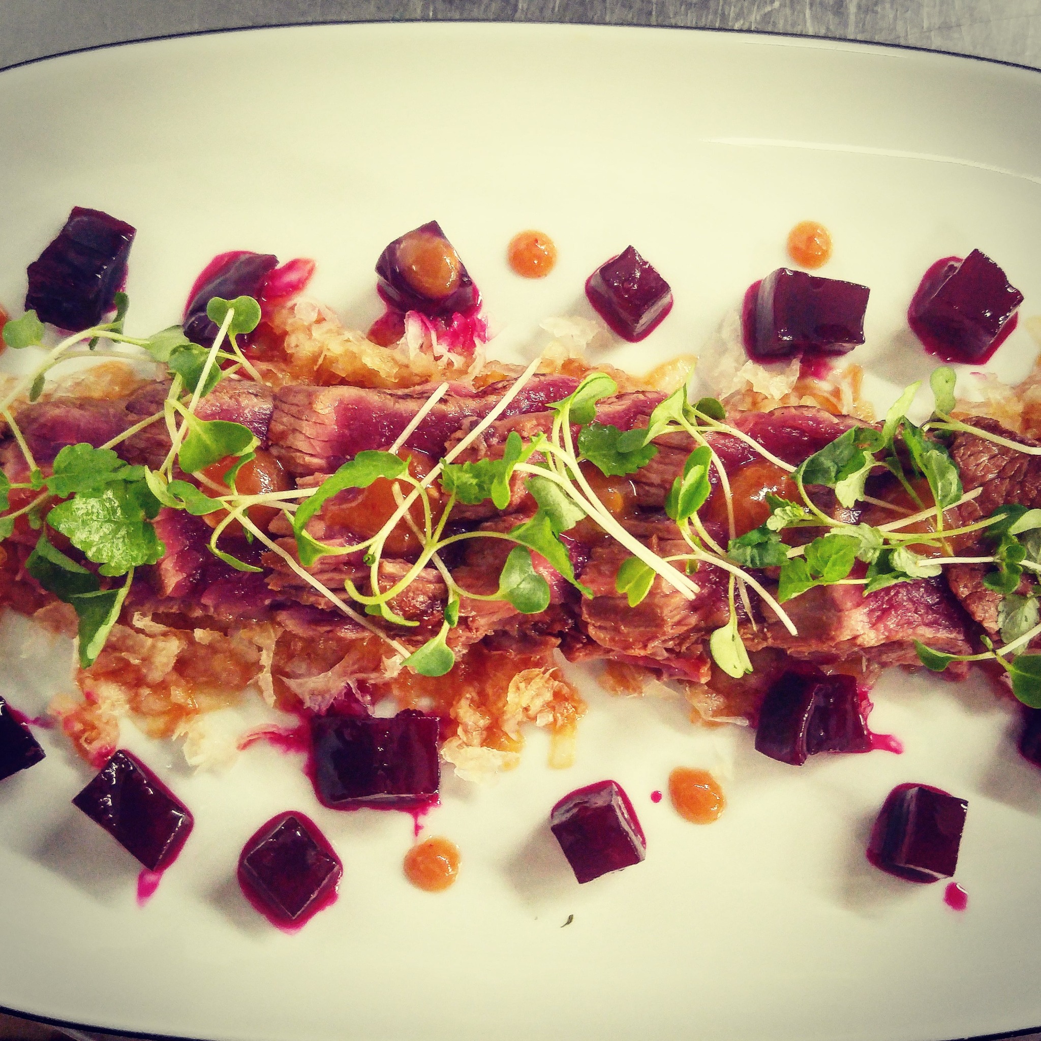 Elegancko podane plastry rostbefu z buraczkami w kostkach, sosem i świeżymi kiełkami na białym talerzu.