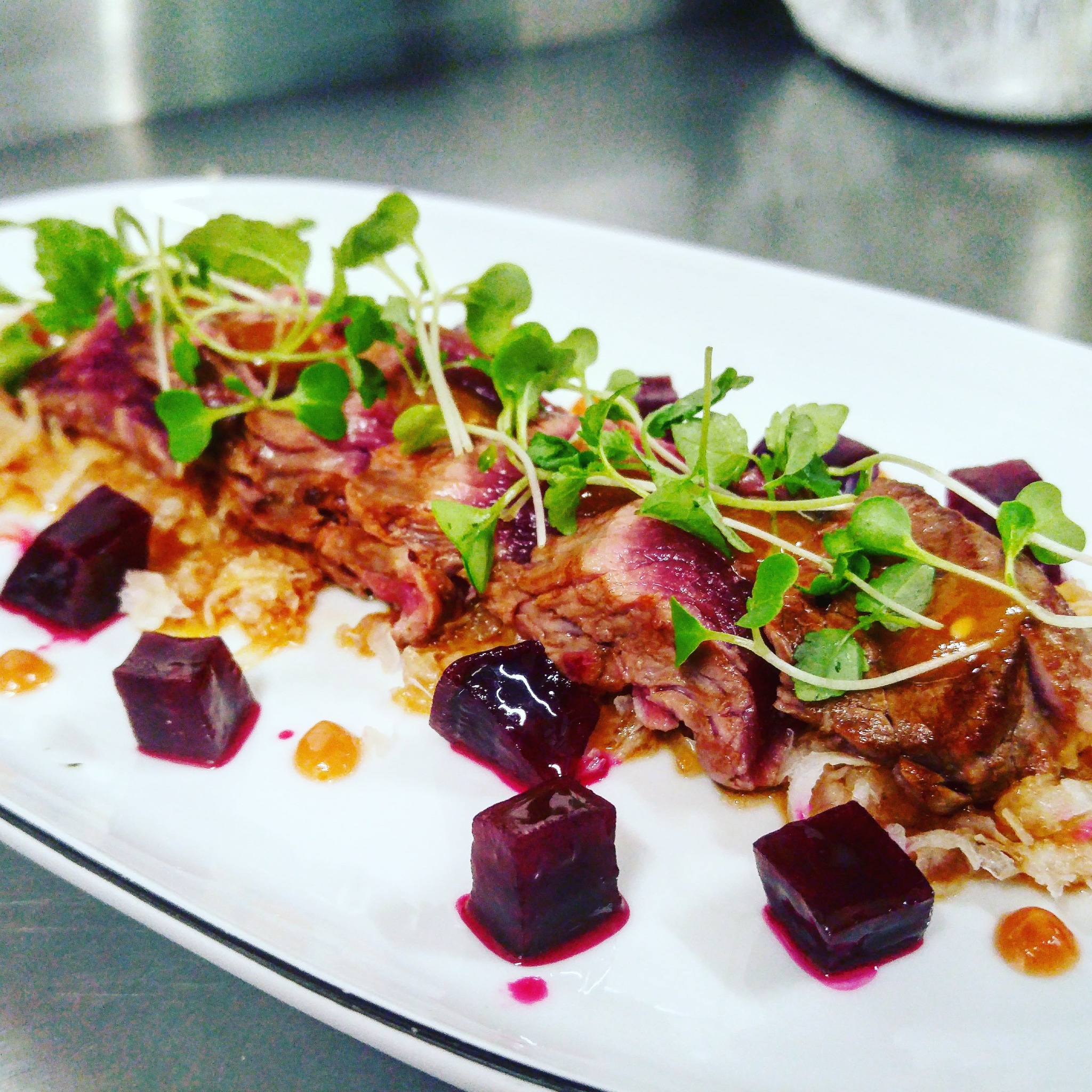 Elegancko podane danie z plastrami wołowiny, kostkami buraka, kiełkami i sosem na białym, prostokątnym talerzu, widoczne w tle stalowe blaty kuchenne.