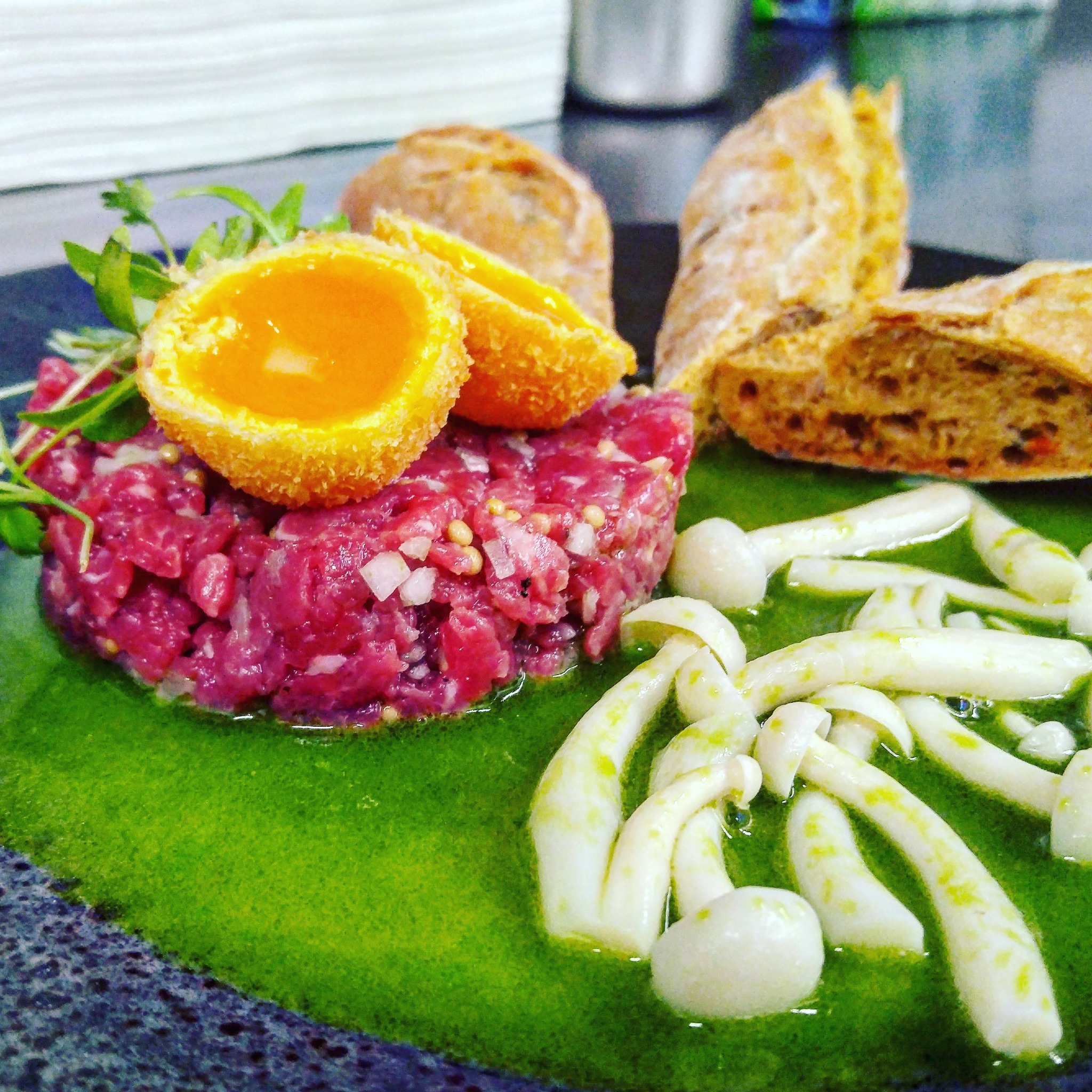 Wykwintny talerz z tatarem wołowym, smażonym jajkiem przepiórczym w panko, pieczywem i marynowanymi grzybami w zielonym sosie.