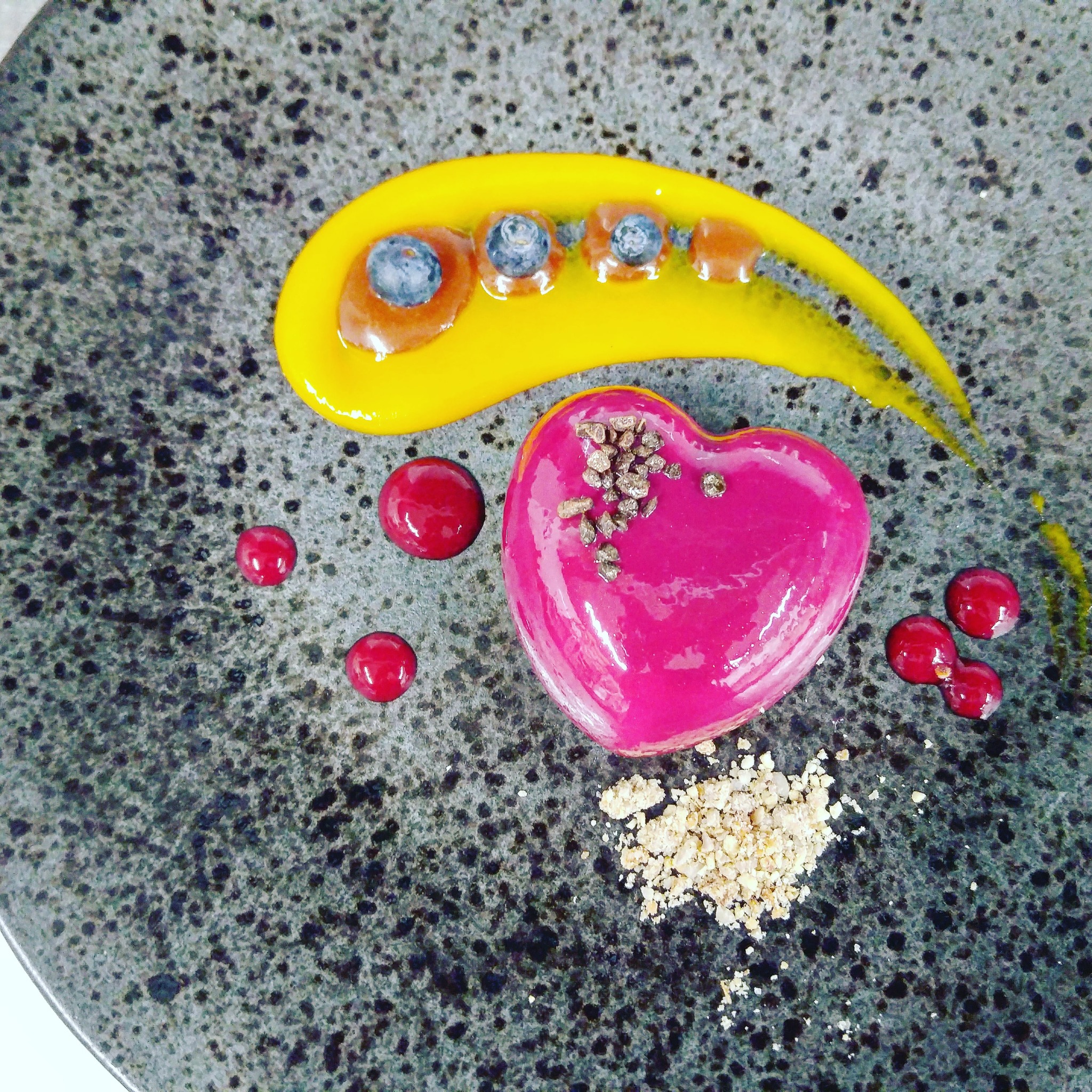 Deser w kształcie serca z różową polewą, posypany kruszonką, otoczony sosem mango z borówkami i czerwonymi kuleczkami na szarym, nakrapianym talerzu.