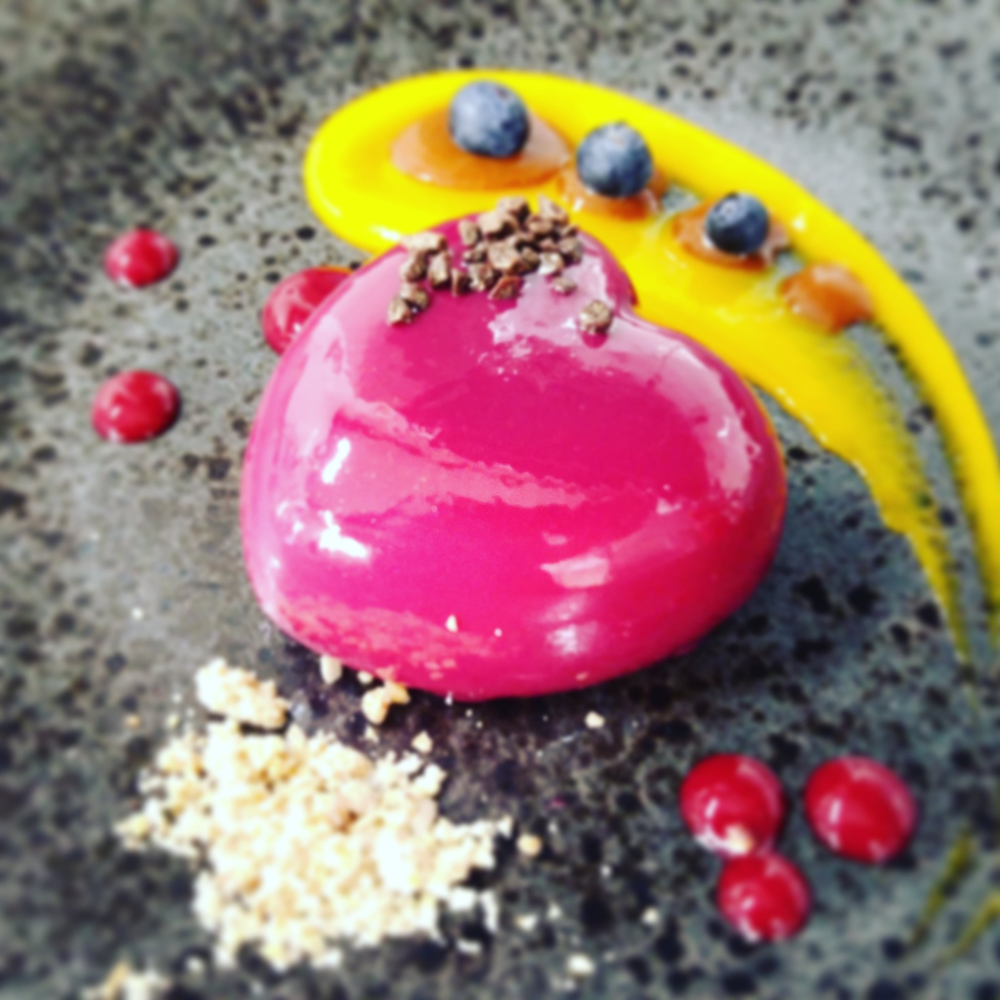 Lśniący, różowy deser w kształcie kopuły, posypany drobnymi kawałkami czekolady, otoczony żółtym sosem z borówkami, czerwonymi żelkami i kruszonką na szarym, teksturowanym tle.