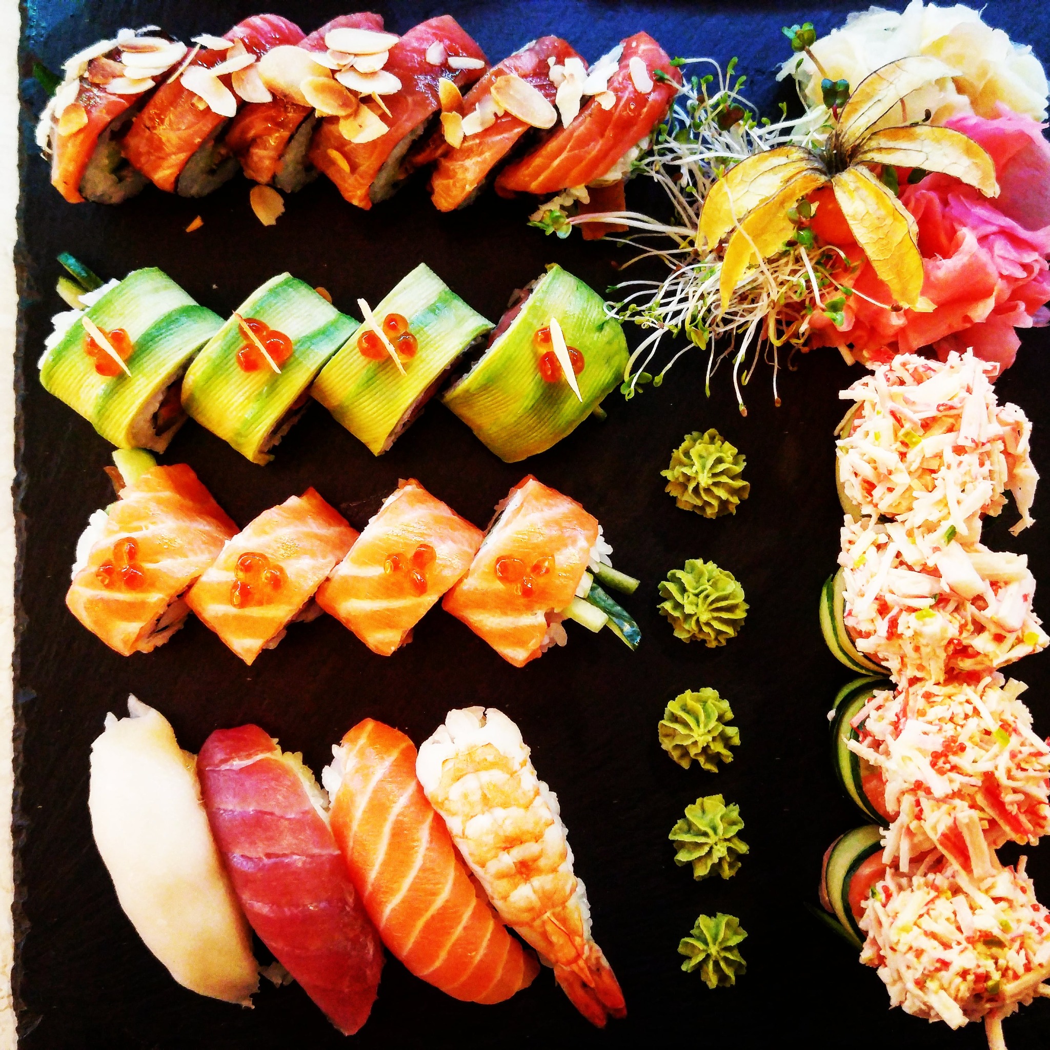 Zestaw sushi z różnymi rodzajami ryb, owoców morza i warzyw, ułożony na czarnym kamiennym talerzu, udekorowany kiełkami i physalis.