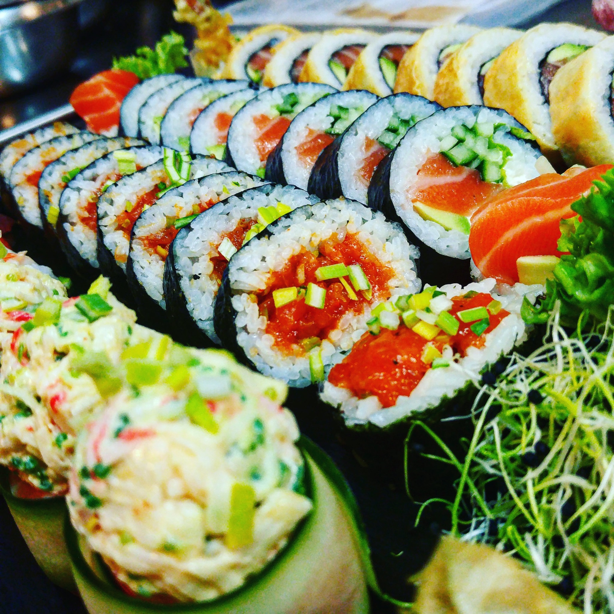 Zestaw sushi z różnymi rodzajami rolek: z łososiem i awokado, z tuńczykiem i posypką z zielonego szczypiorku, oraz sałatka z krabowych paluszków i kiełki, ułożone na czarnym talerzu.