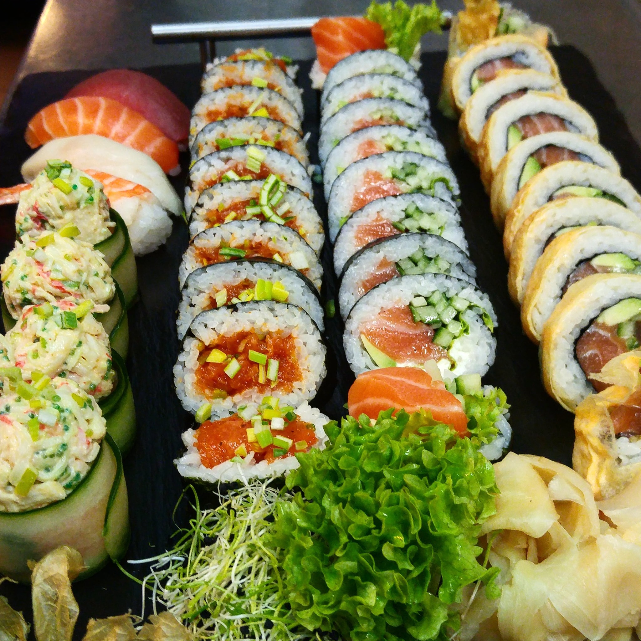 Różnorodny zestaw sushi na czarnym talerzu: nigiri z łososiem i tuńczykiem, california rolls z awokado i łososiem, maki z ogórkiem i pastą z tuńczyka, dekorowane zielonymi pędami i sałatą,...