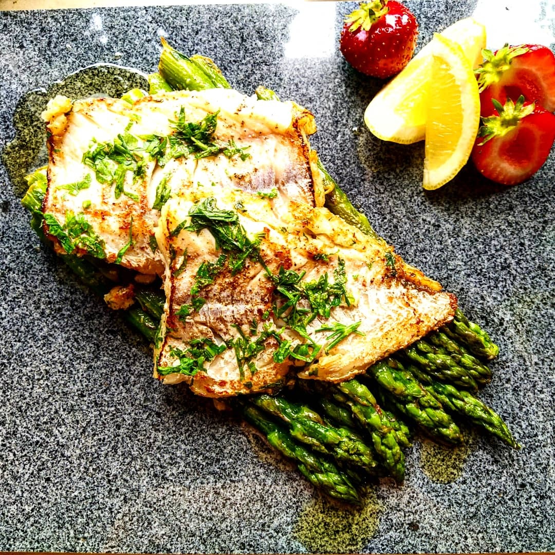 Filet rybny z grilla posypany ziołami, ułożony na szparagach, z dodatkiem truskawek i cytryny na szarym kamiennym talerzu.
