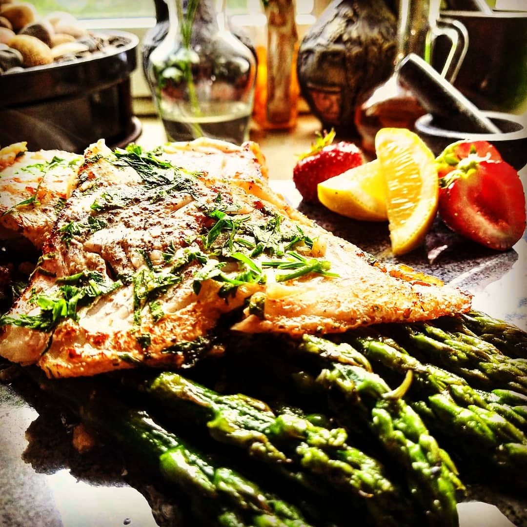 Filet rybny posypany ziołami, ułożony na szparagach, z dodatkiem truskawek i cytryny, w tle moździerz i inne naczynia.