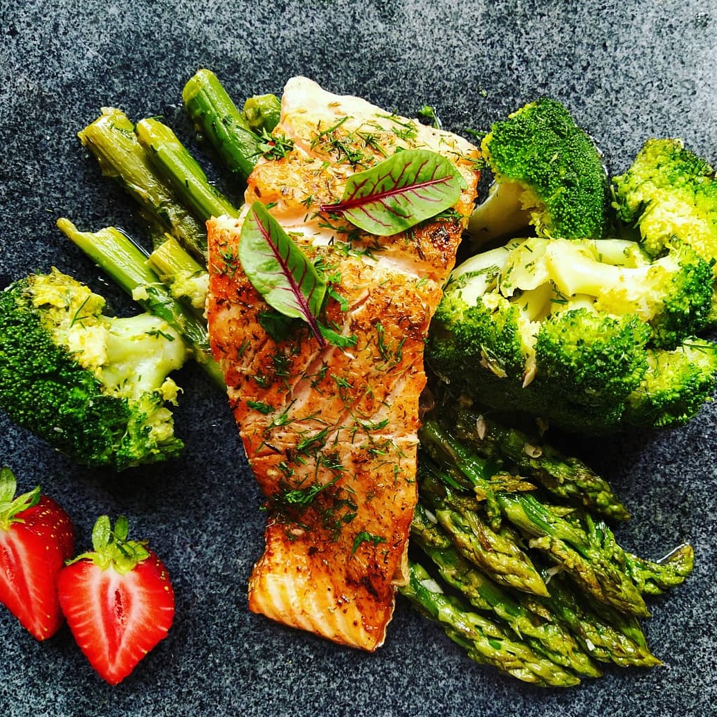 Grillowany filet z łososia posypany koperkiem, podany z brokułami, szparagami i dwoma połówkami truskawek na ciemnym, kamiennym talerzu.
