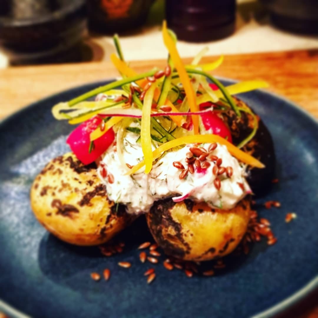 Opiekane ziemniaki z kremowym serem, rzodkiewką, ogórkiem, marchewką i posypką z siemienia lnianego na ciemnoniebieskim talerzu.