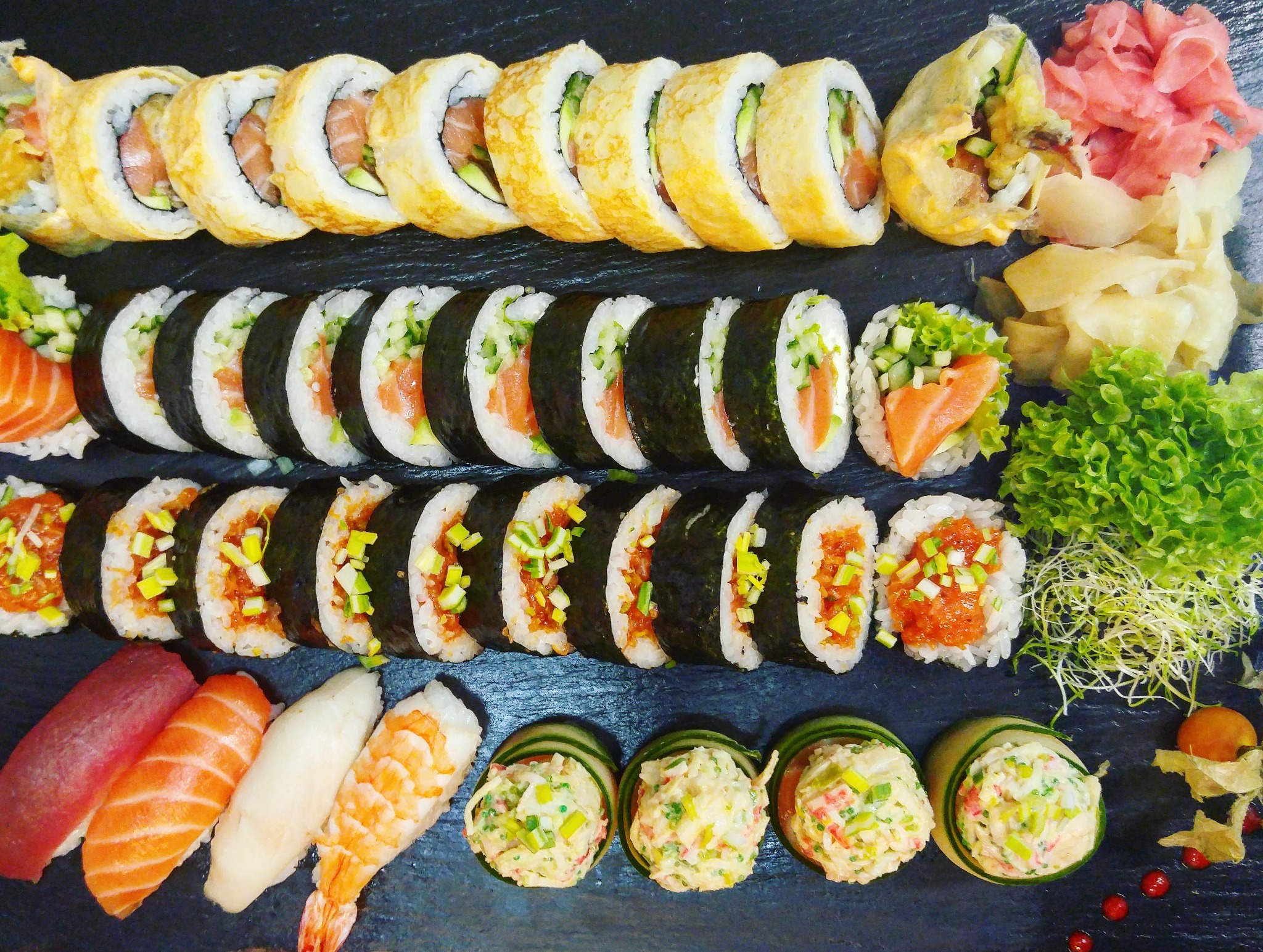 Zestaw sushi: rolki z łososiem i ogórkiem zawinięte w nori, rolki w tempurze, nigiri z tuńczykiem i krewetką, sałatka z paluszków krabowych w plastrach ogórka, imbir i sałata na czarnym tle.