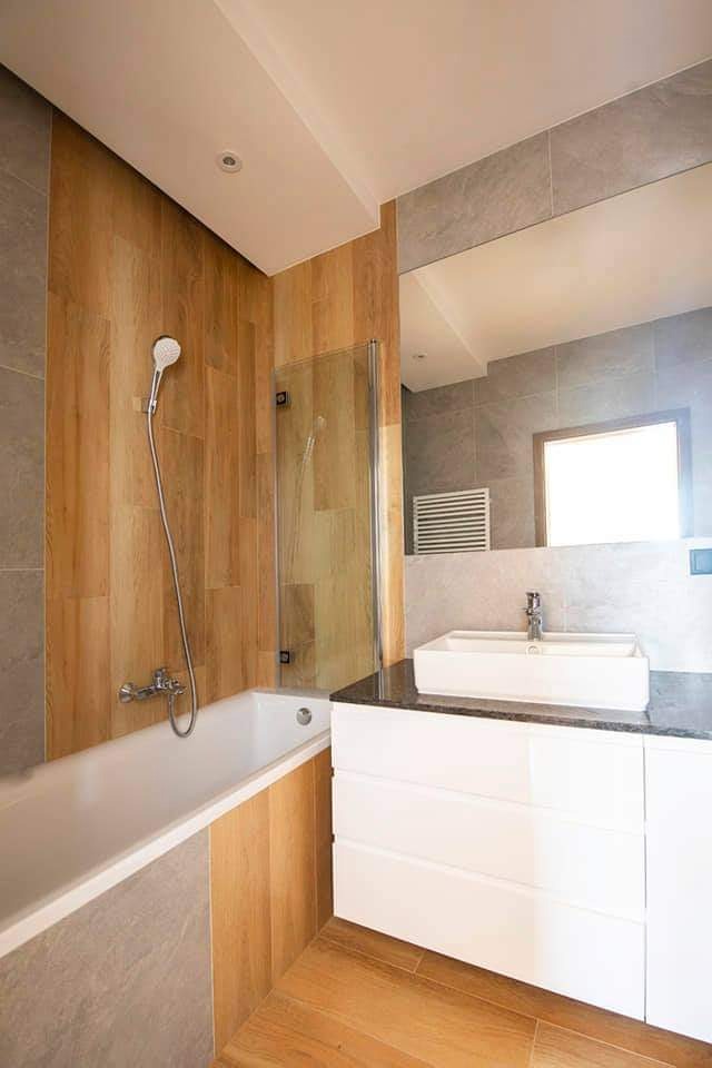 Nowoczesna łazienka z drewnopodobnymi płytkami na ścianie przy wannie i szarą glazurą na pozostałych ścianach, biała umywalka nablatowa z czarnym blatem i lustrem.