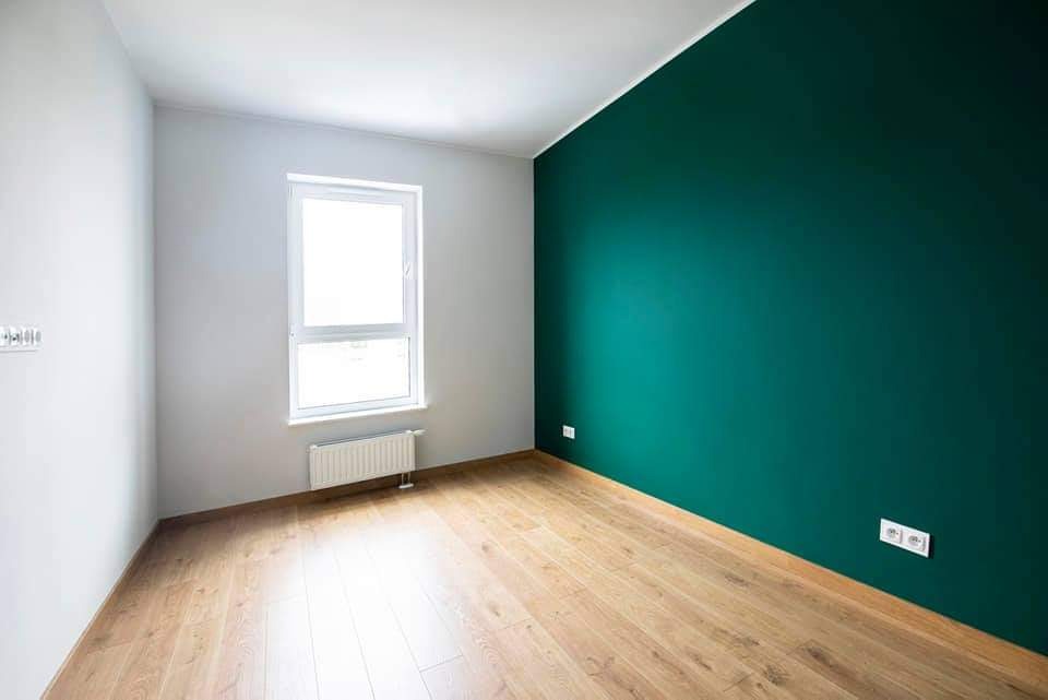 Pomalowany na biało pokój z drewnianą podłogą i ścianą w kolorze ciemnej zieleni, okno z grzejnikiem pod spodem.