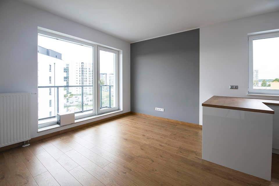 Jasne wnętrze pokoju z drewnianą podłogą, szarą ścianą akcentującą i dużym oknem z widokiem na miasto.